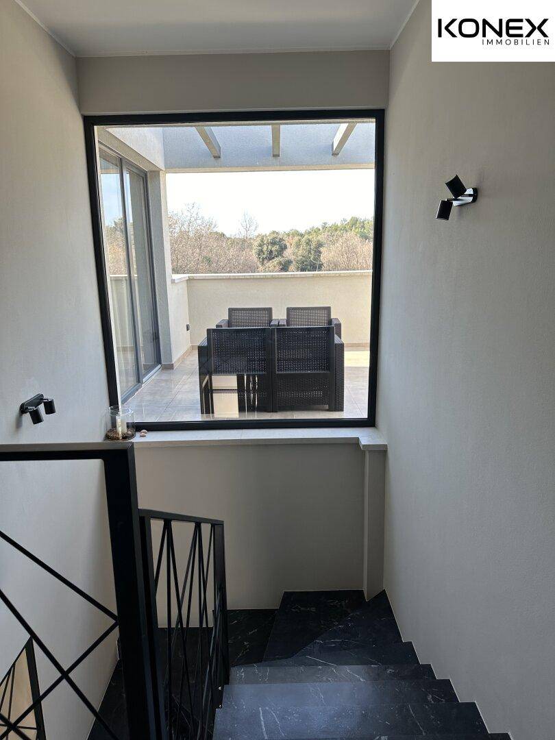 Treppenaufgang mit Blick auf eine möblierte Dachterrasse durch große Fenster.