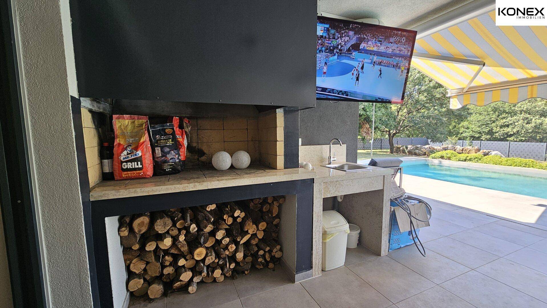 Funktionale Außenküche mit Grillbereich, Spüle und Holzvorrat neben dem Pool.