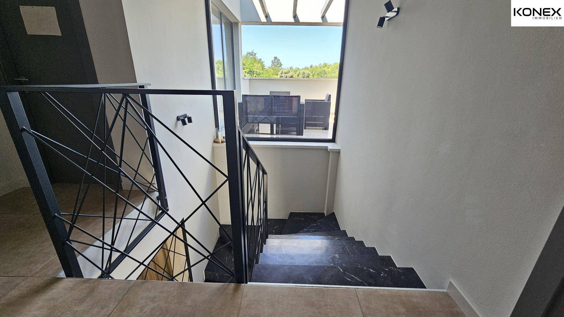Treppenaufgang mit Blick auf eine möblierte Dachterrasse durch große Fenster.
