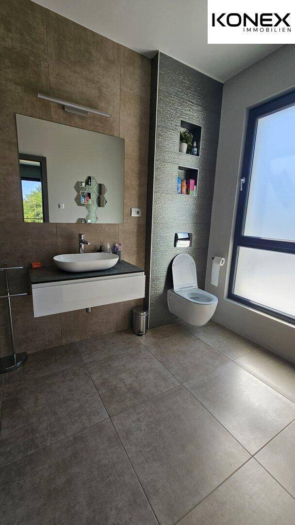 Helles Badezimmer mit ebenerdiger Dusche, Waschbecken, Wand-WC und Fenster.