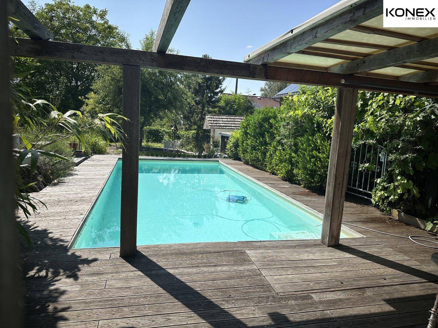 Swimmingpool mit Holzdeck, gesehen durch eine Holzpergola, umgeben von Bäumen.
