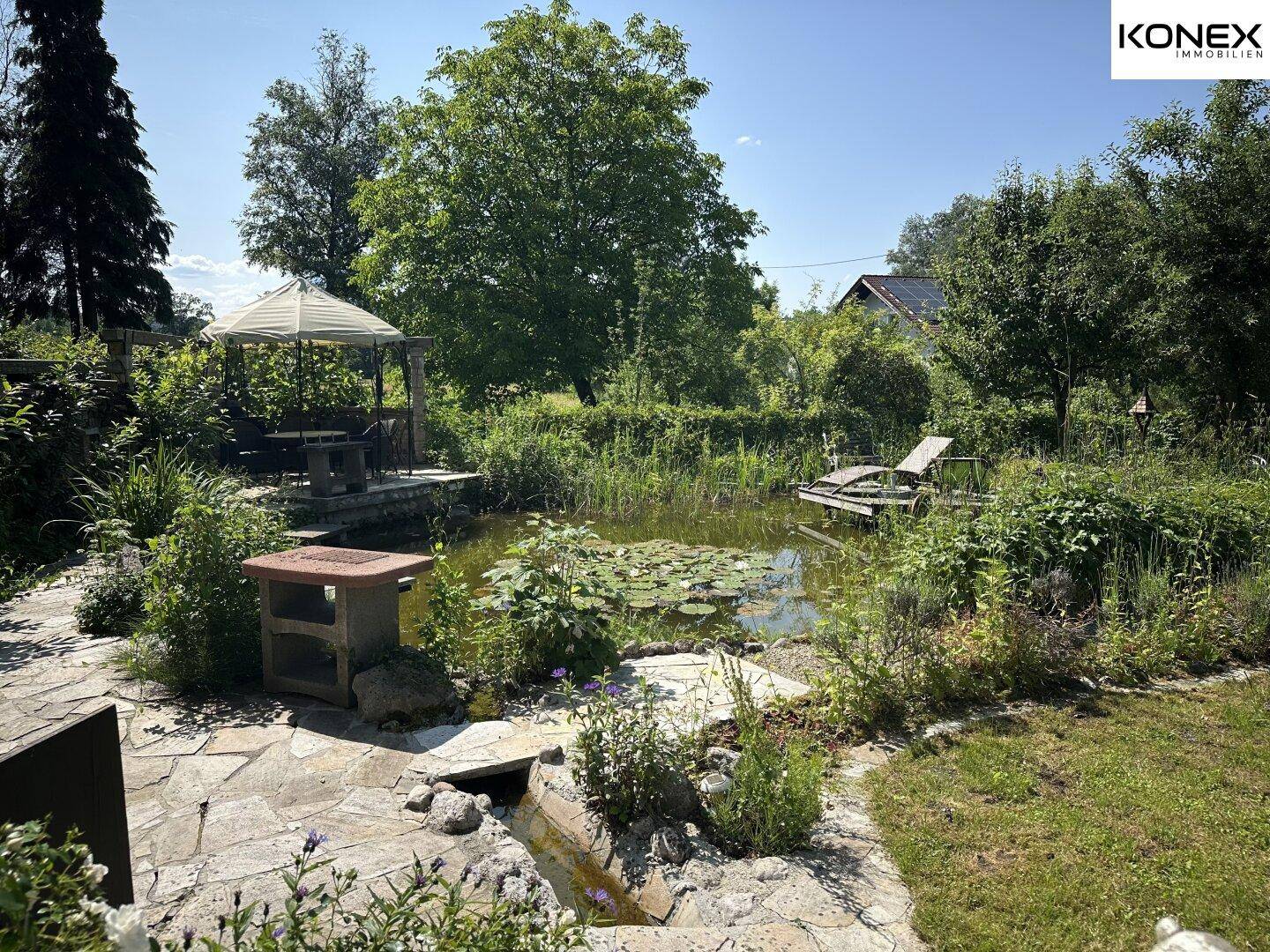 Idyllischer Gartenbereich mit Teich, Steinterrasse und einem schattenspendenden Pavillon.