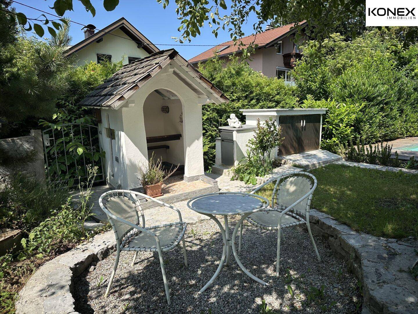 Kleiner Sitzbereich im Garten mit einem dekorativen Schrein und üppiger Bepflanzung.