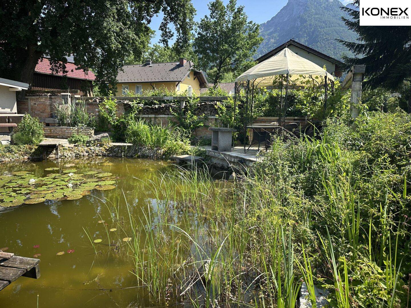 Naturnaher Gartenteich mit üppiger Bepflanzung und einem Pavillon im Hintergrund.