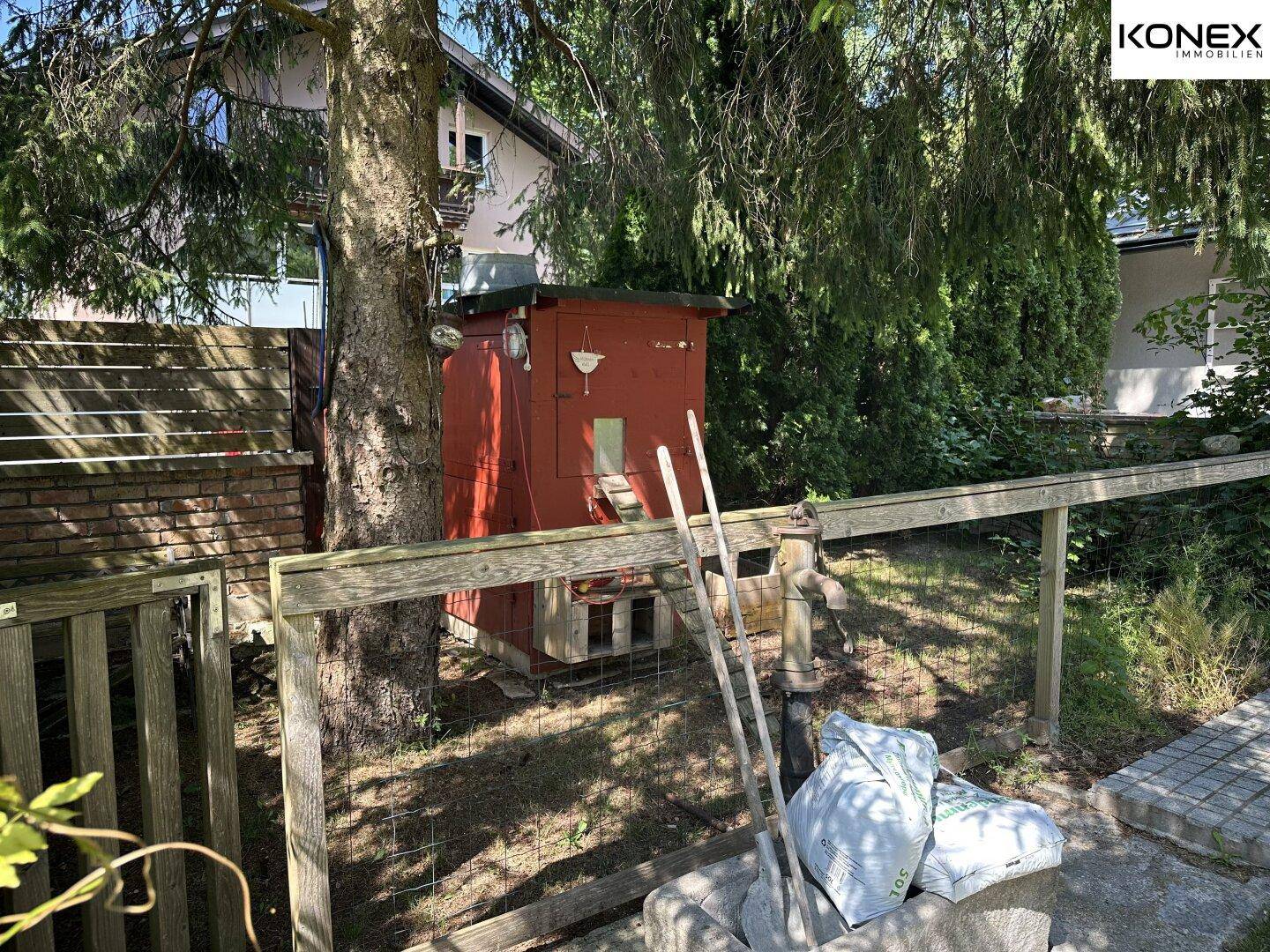 Roter Gartenschuppen umgeben von Bäumen und einem Holzzaun im sonnigen Garten.