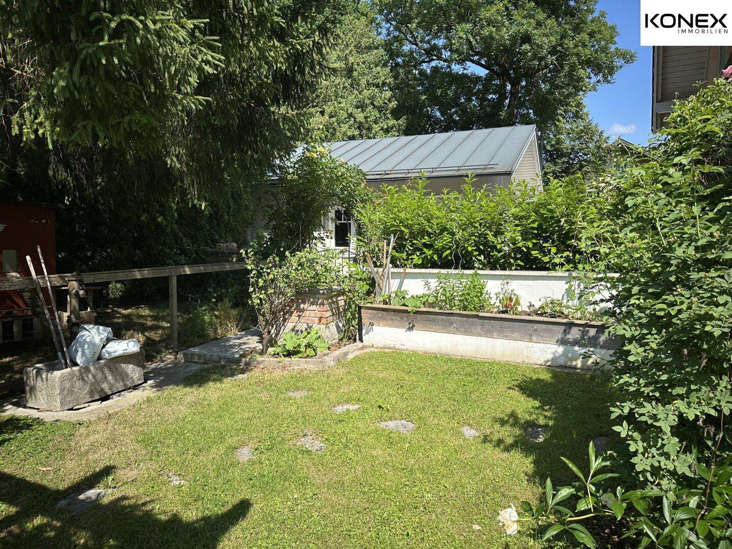 Gartenbereich mit Hochbeet und einem kleinen Schuppen im Hintergrund, umgeben von Grün.