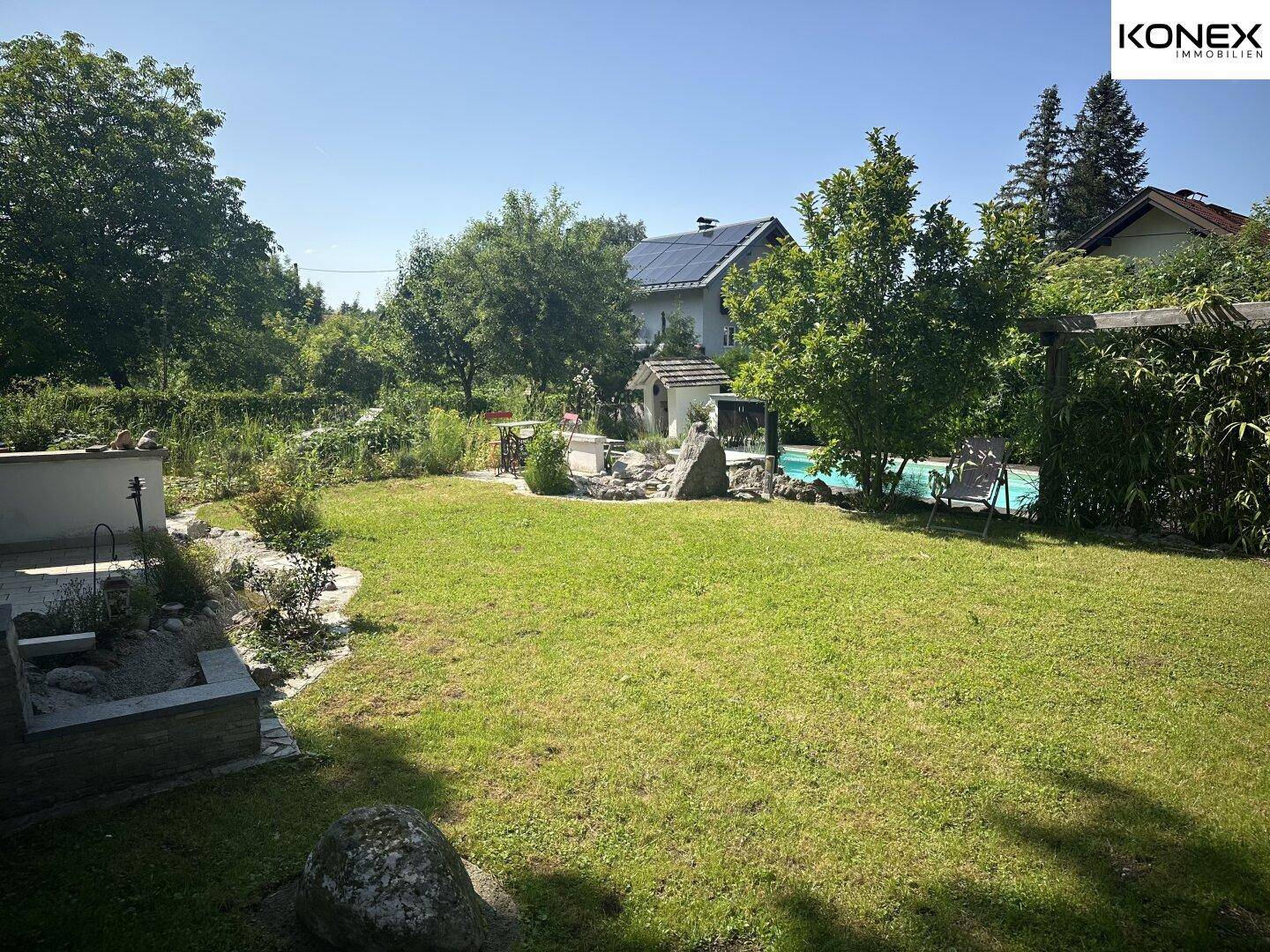 Weitläufiger Garten mit Rasenfläche, Pool und Haus im Hintergrund unter blauem Himmel.