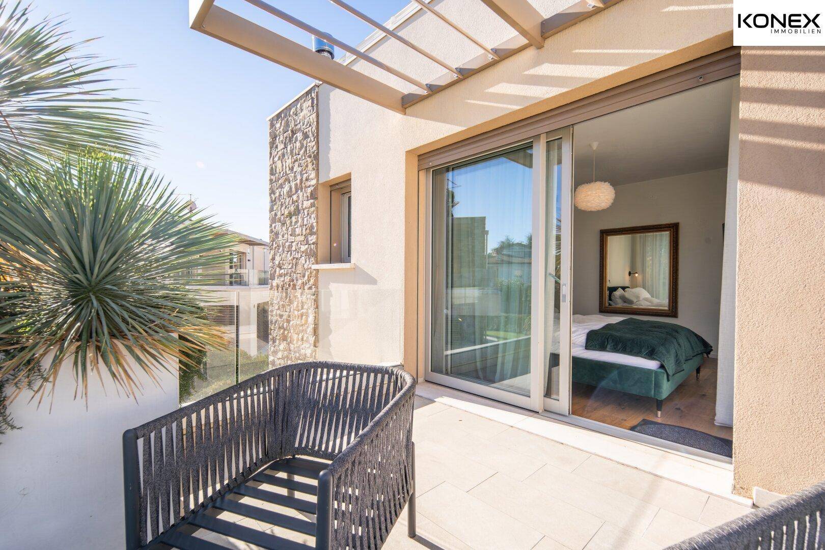 Sonniger Balkon mit modernen Sitzgelegenheiten und Zugang zu einem Schlafzimmer, umgeben von Pflanzen.