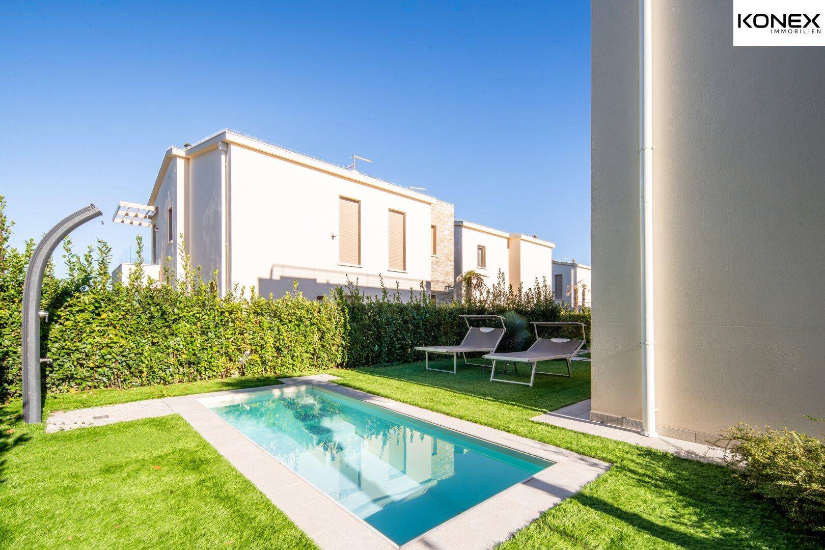 Sonniger Gartenbereich mit kleinem Pool, Außendusche und Liegestühlen, umgeben von Hecken.