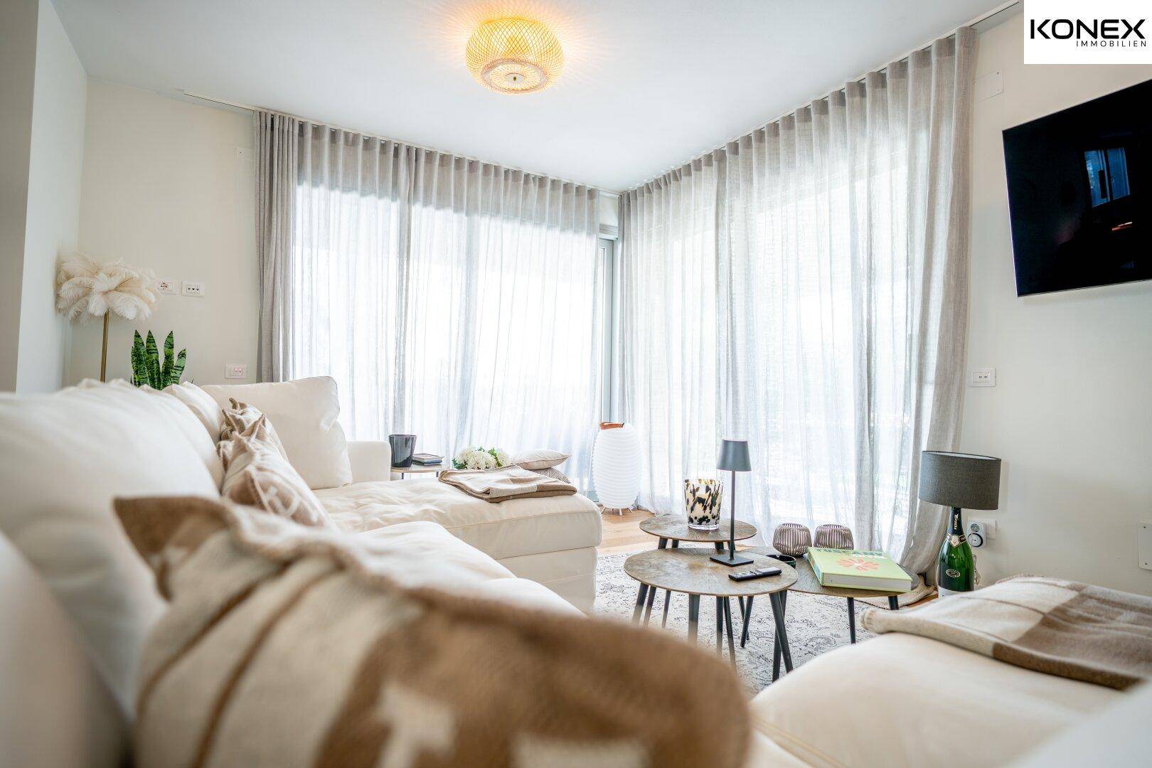Gemütliches Wohnzimmer mit großzügigem Sofa, hellen Vorhängen und viel Tageslicht durch große Fenster.