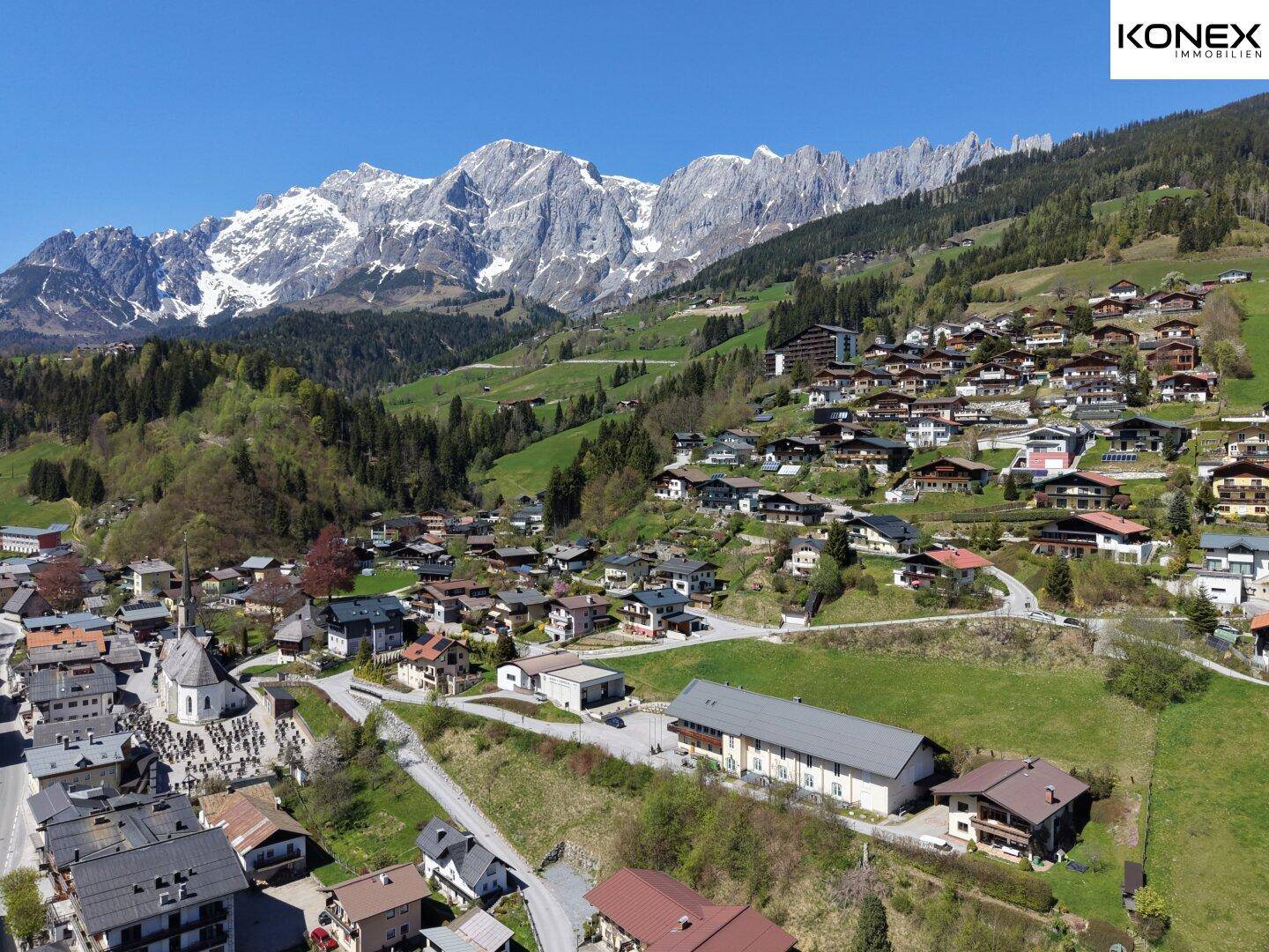 Luftaufnahme eines charmanten Bergdorfes, eingebettet in grüne Hügel mit majestätischen Bergen im Hintergrund.