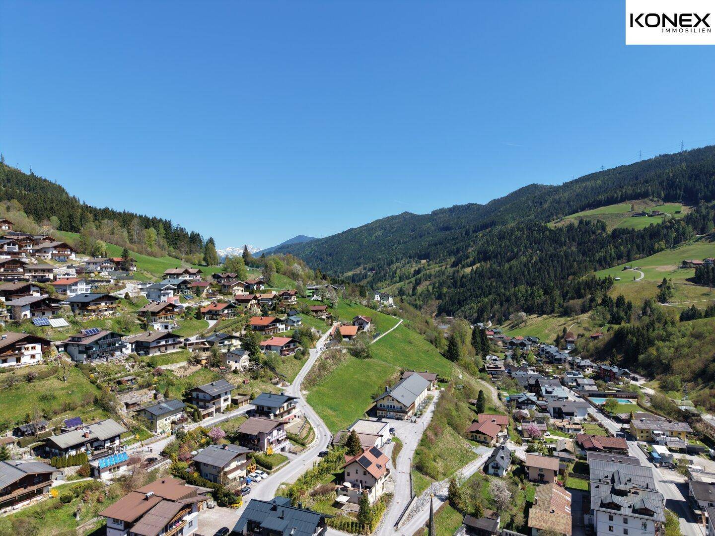 Weitläufige Luftaufnahme eines Bergdorfes in einem grünen Tal, umgeben von bewaldeten Hängen unter klarem Himmel.