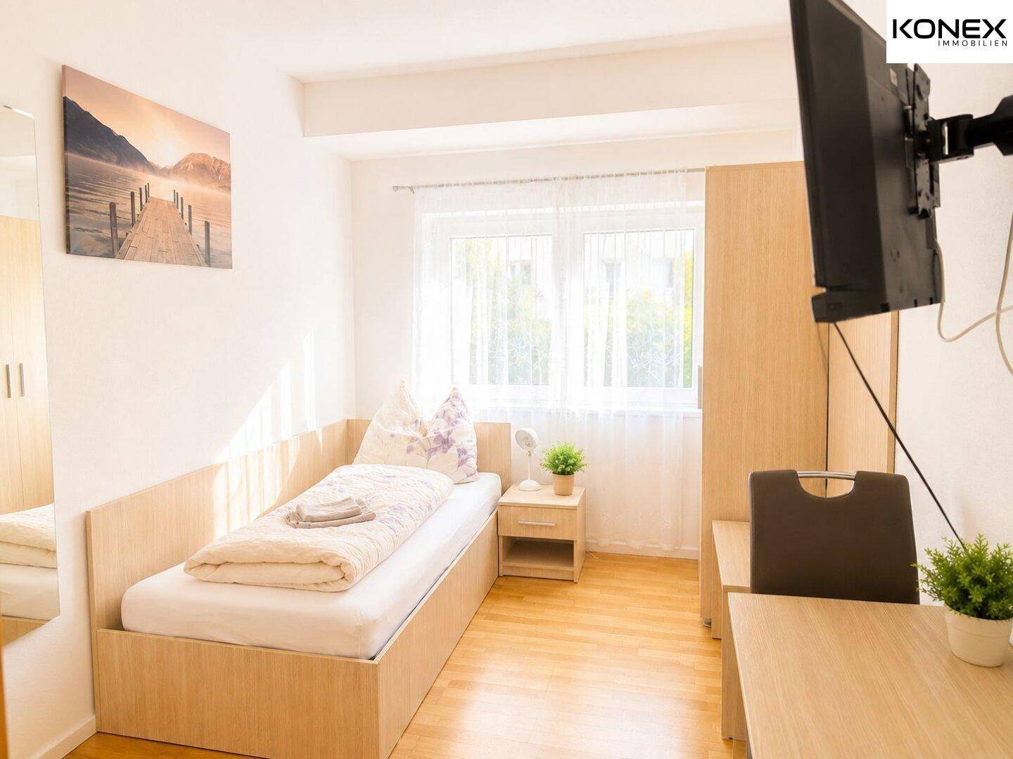 Einzelzimmer mit Bett, Schreibtisch, Wandfernseher und hellem Holzboden.