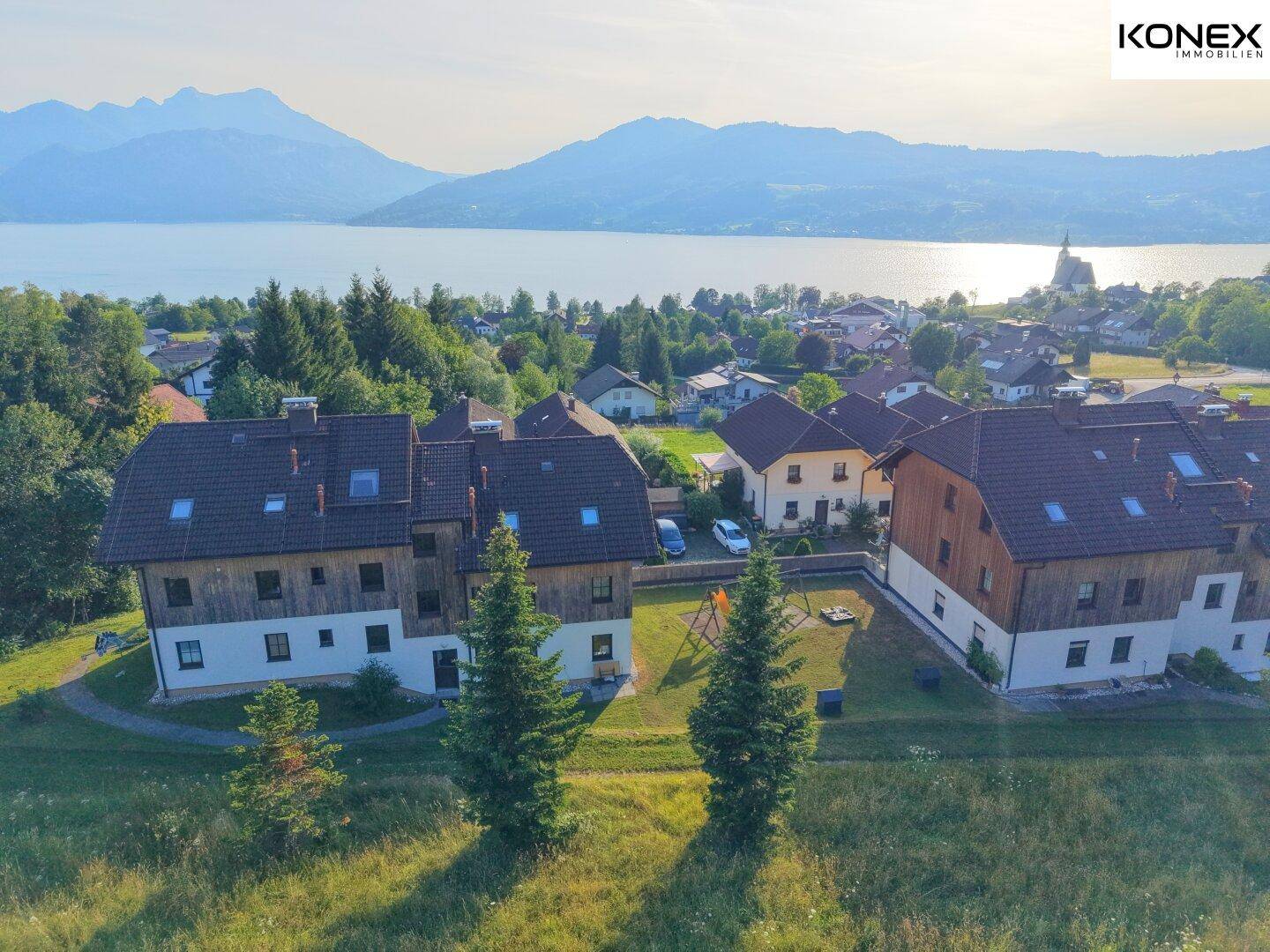 Weitläufige Ansicht der Wohnanlage mit See, Bergen und einem Spielbereich für Kinder.