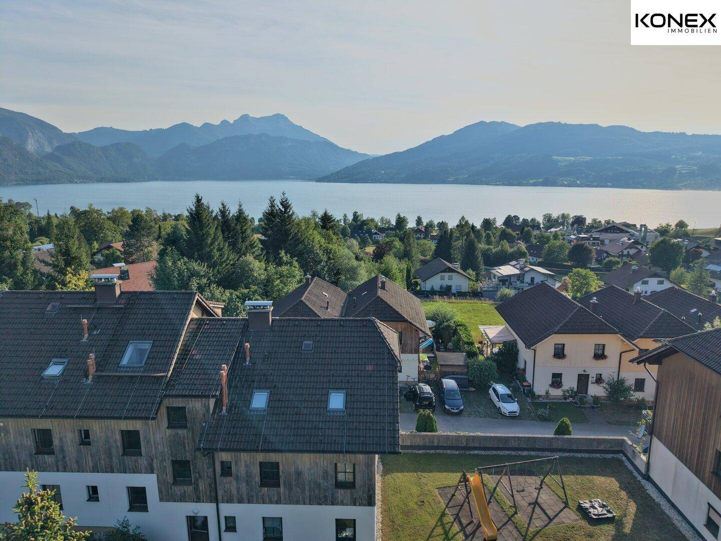 Panoramablick auf die Wohnanlage mit See, Bergen und einem Spielplatz im Vordergrund.