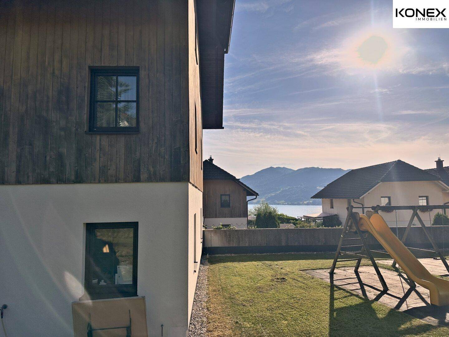 Seitlicher Blick auf das Gebäude mit Rasenfläche, Spielplatz und See- sowie Bergblick.