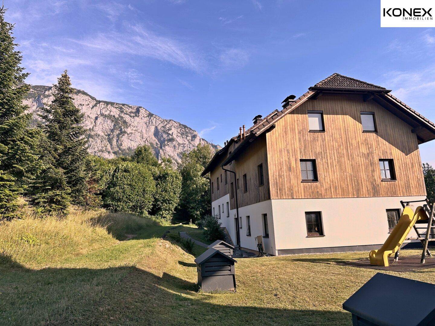 Gebäudeansicht mit angrenzendem Rasen, Spielplatz und beeindruckendem Bergpanorama.