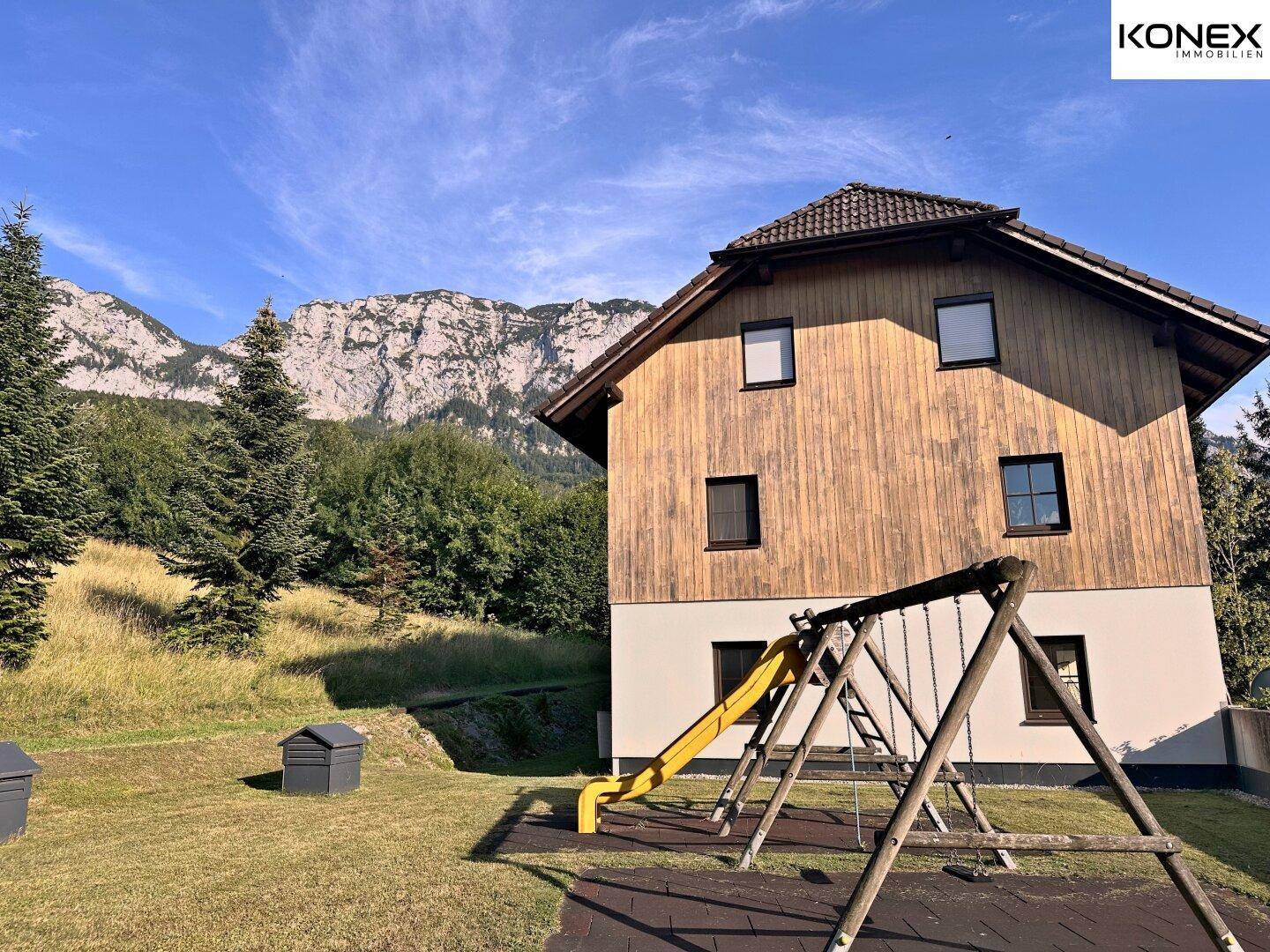Seitenansicht des Hauses mit Spielplatz auf der Rasenfläche und majestätischen Bergen im Hintergrund.