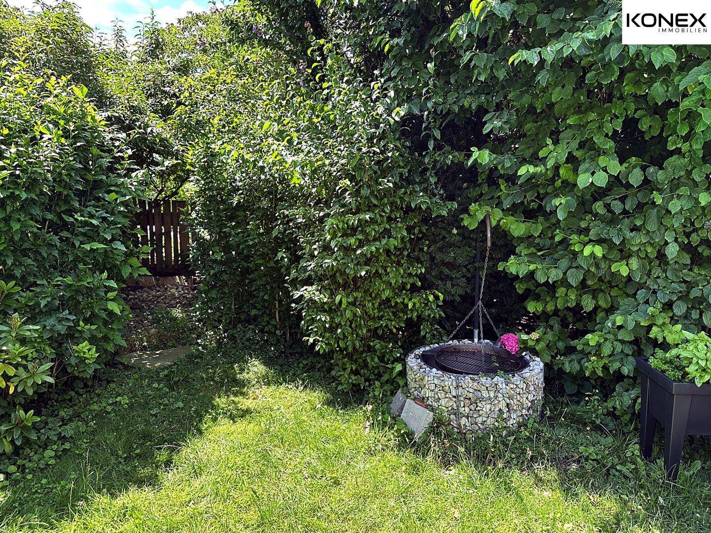Gartenbereich mit einer gemauerten Feuerstelle, umgeben von dichtem Grün.