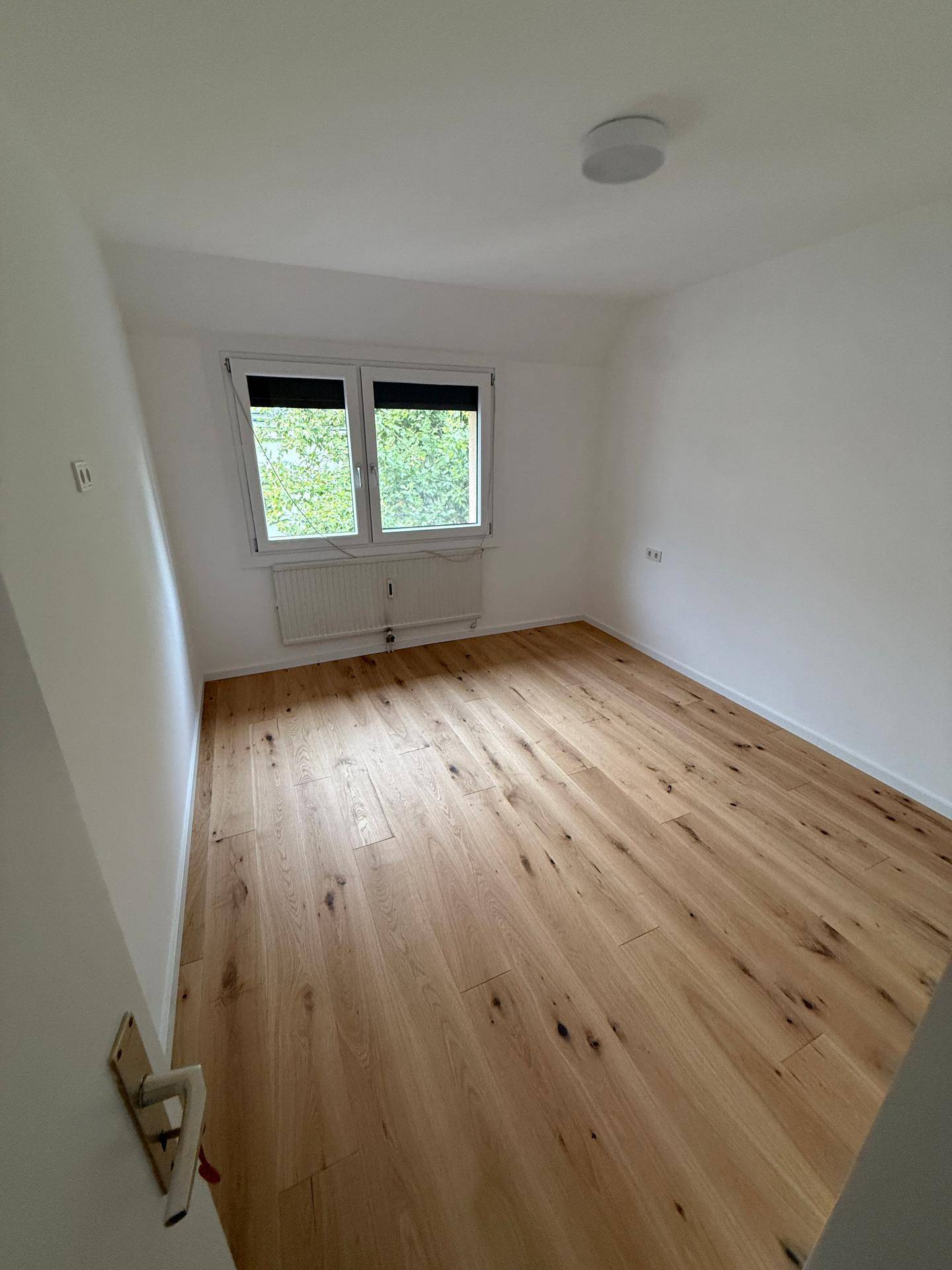 Helles, leeres Zimmer mit Holzboden und Fenster mit Blick ins Grüne.