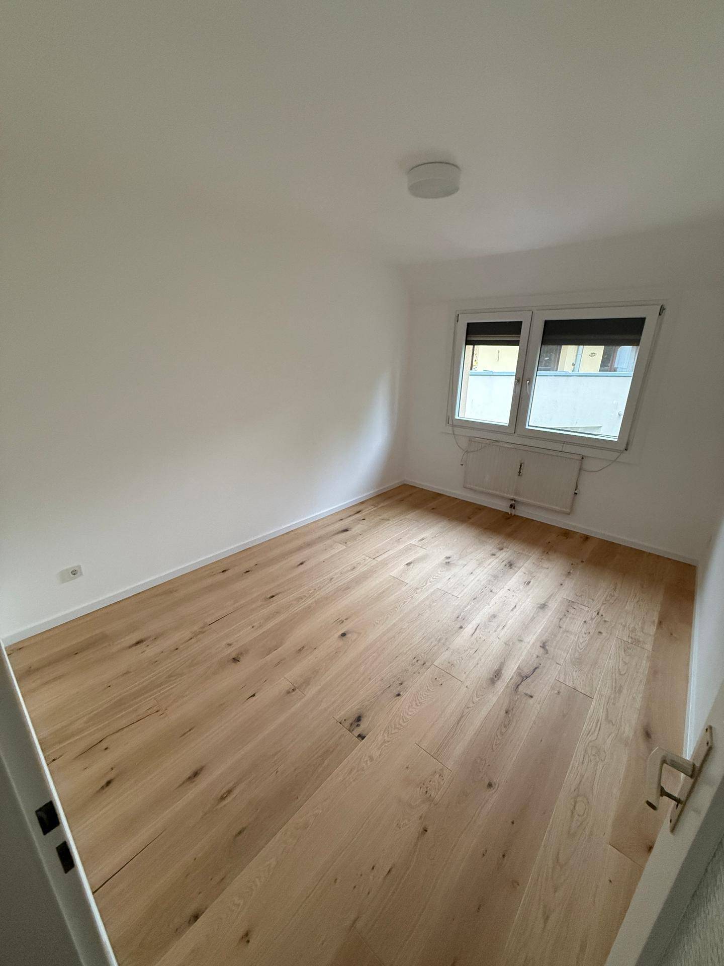 Leeres Zimmer mit hellem Holzboden und Fenster, das viel Tageslicht hereinlässt.