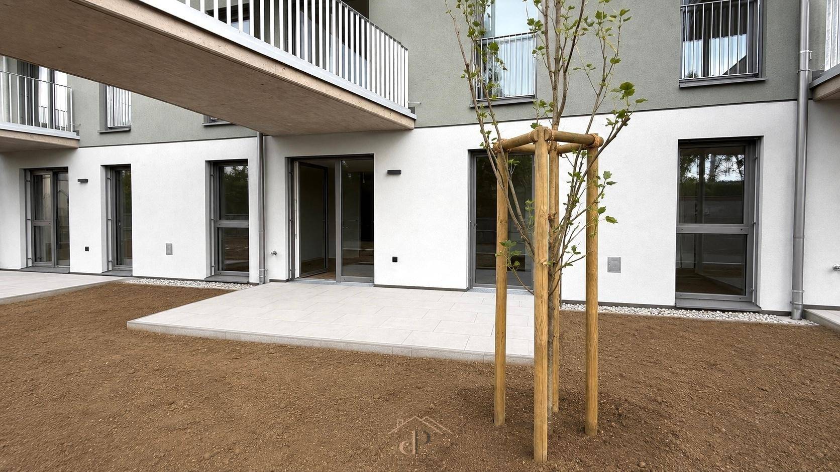 Erdgeschossterrasse eines Neubaus mit jungem Baum und Zugang zum Gartenbereich.