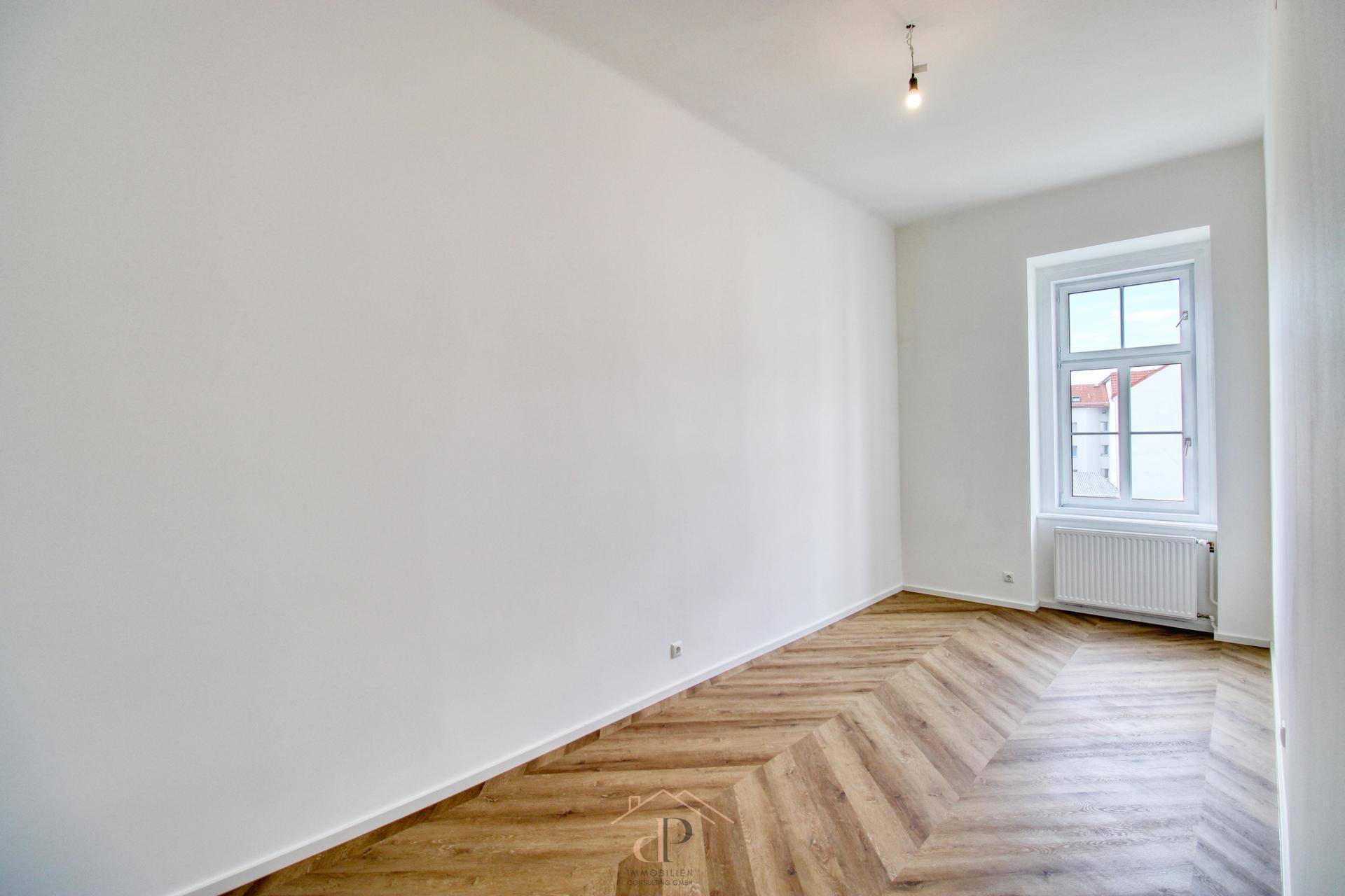 Geräumiger, heller Raum mit klassischem Fischgrätparkett und einem Fenster mit Ausblick.