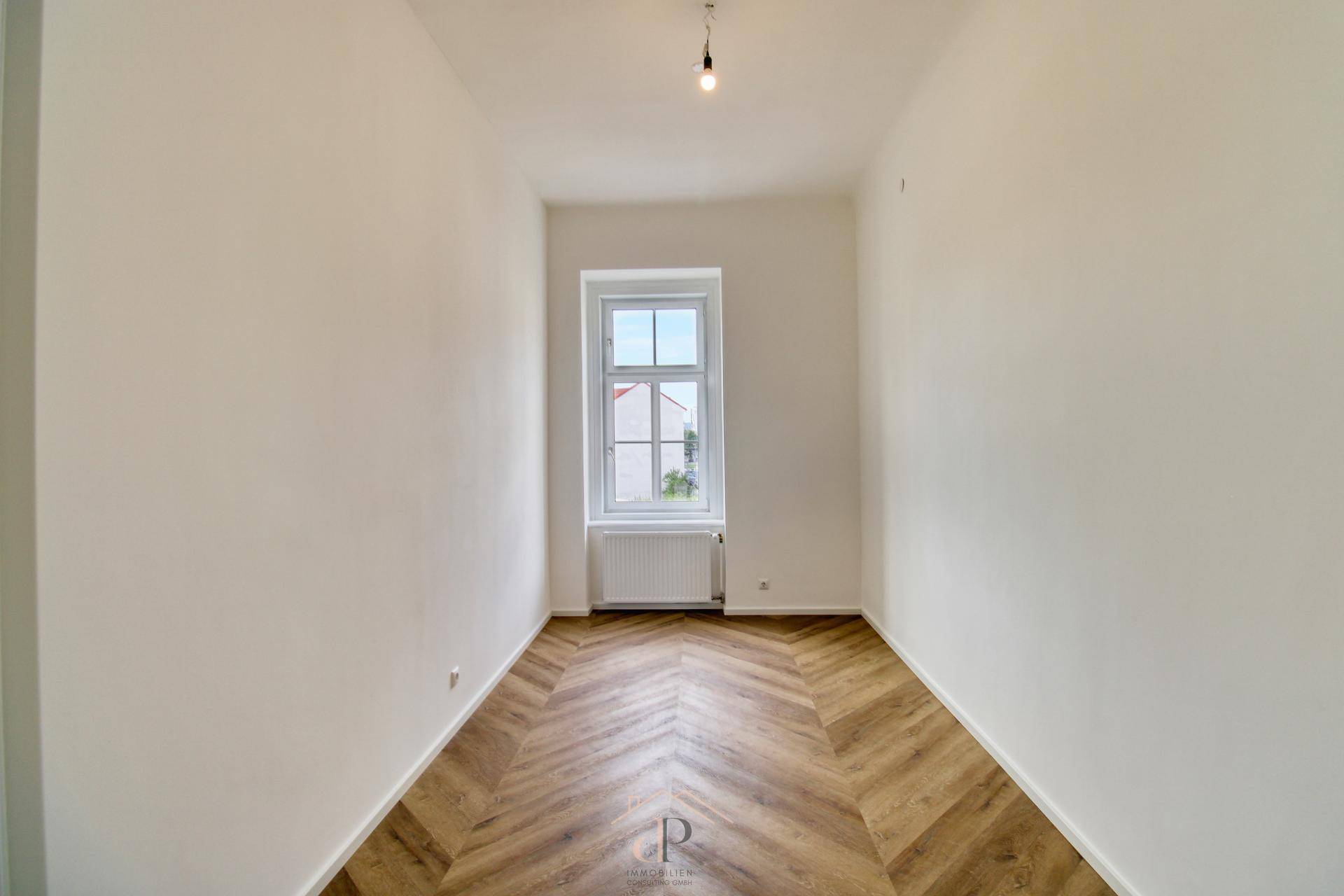 Heller, leerer Raum mit Fischgrätparkett und einem Fenster, das viel Tageslicht hereinlässt.