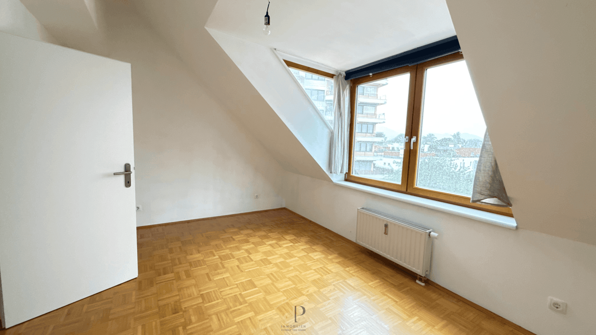 Leeres Schlafzimmer im Dachgeschoss mit schrägen Wänden, großem Fenster und Parkettboden.