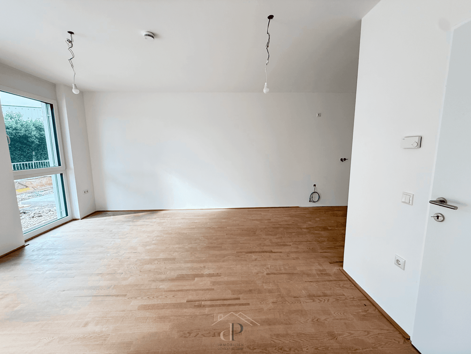 Leerer Raum mit Holzboden und großer Fensterfront, die viel Tageslicht hereinlässt.