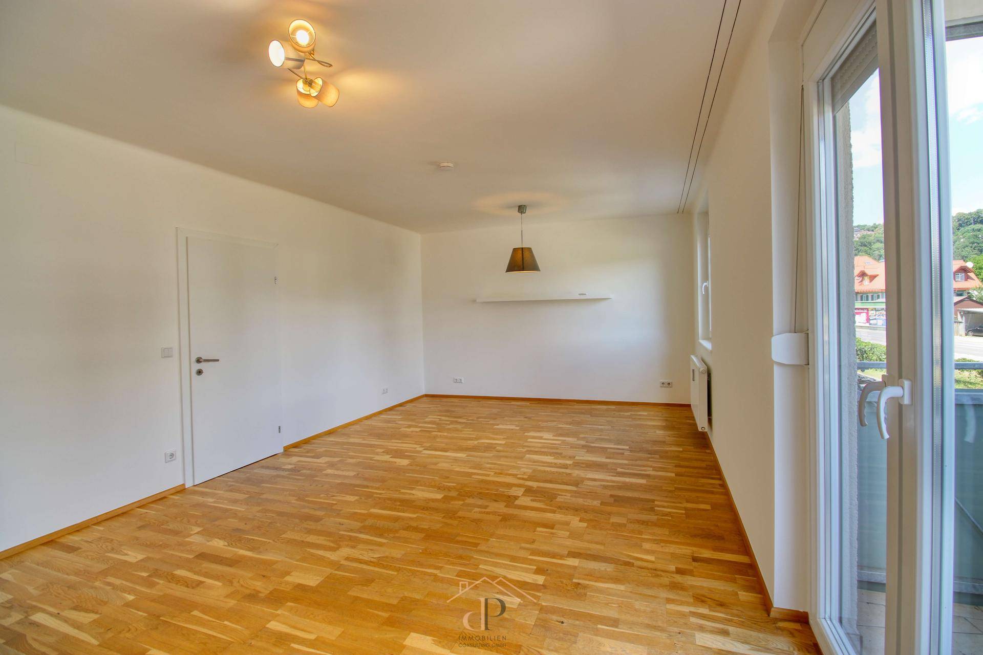 Leeres Wohnzimmer mit Holzboden, weißen Wänden und großem Fenster.
