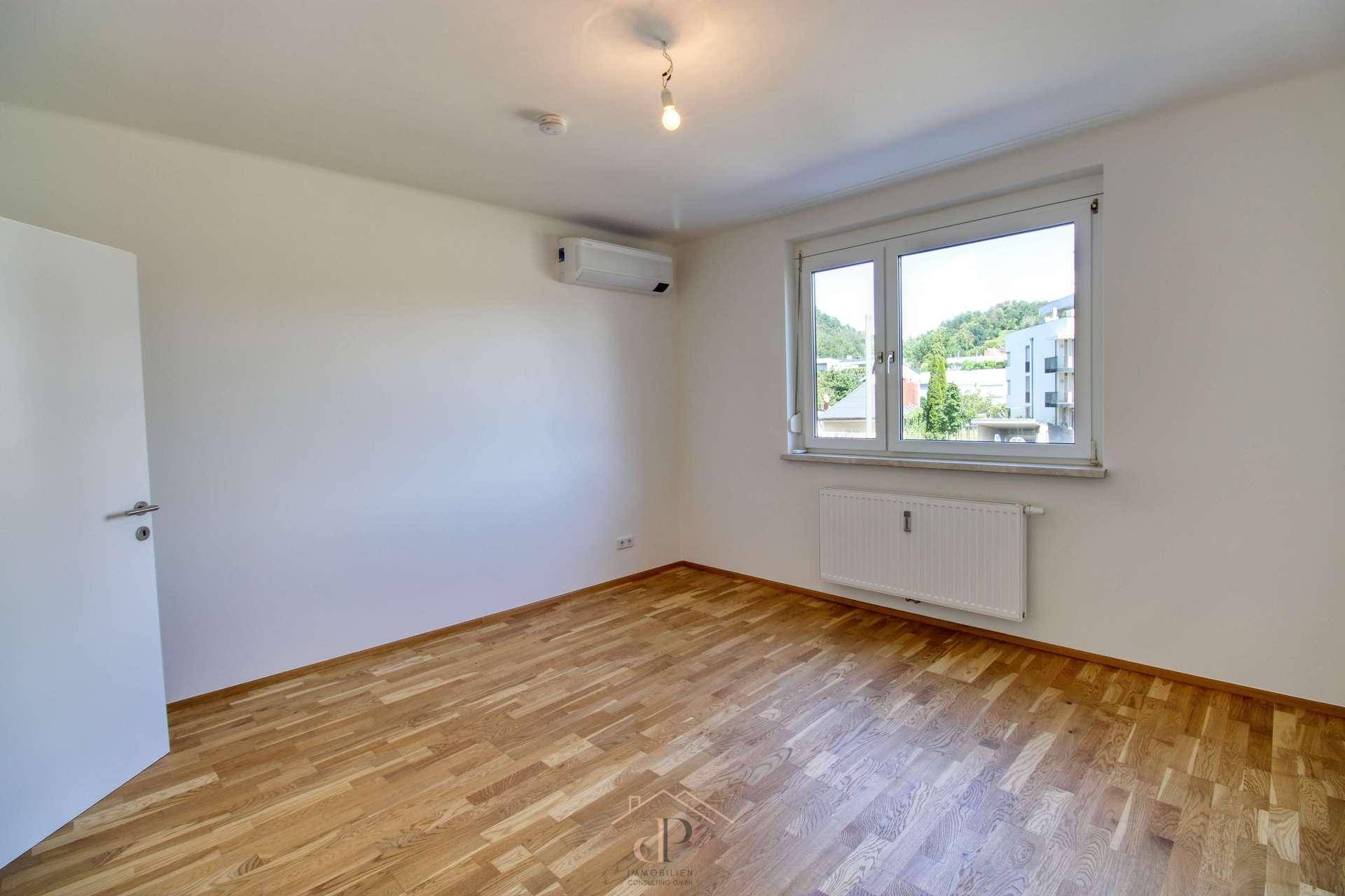 Helles Schlafzimmer mit Holzboden, Fenster und Klimaanlage, bietet einen Blick ins Grüne.