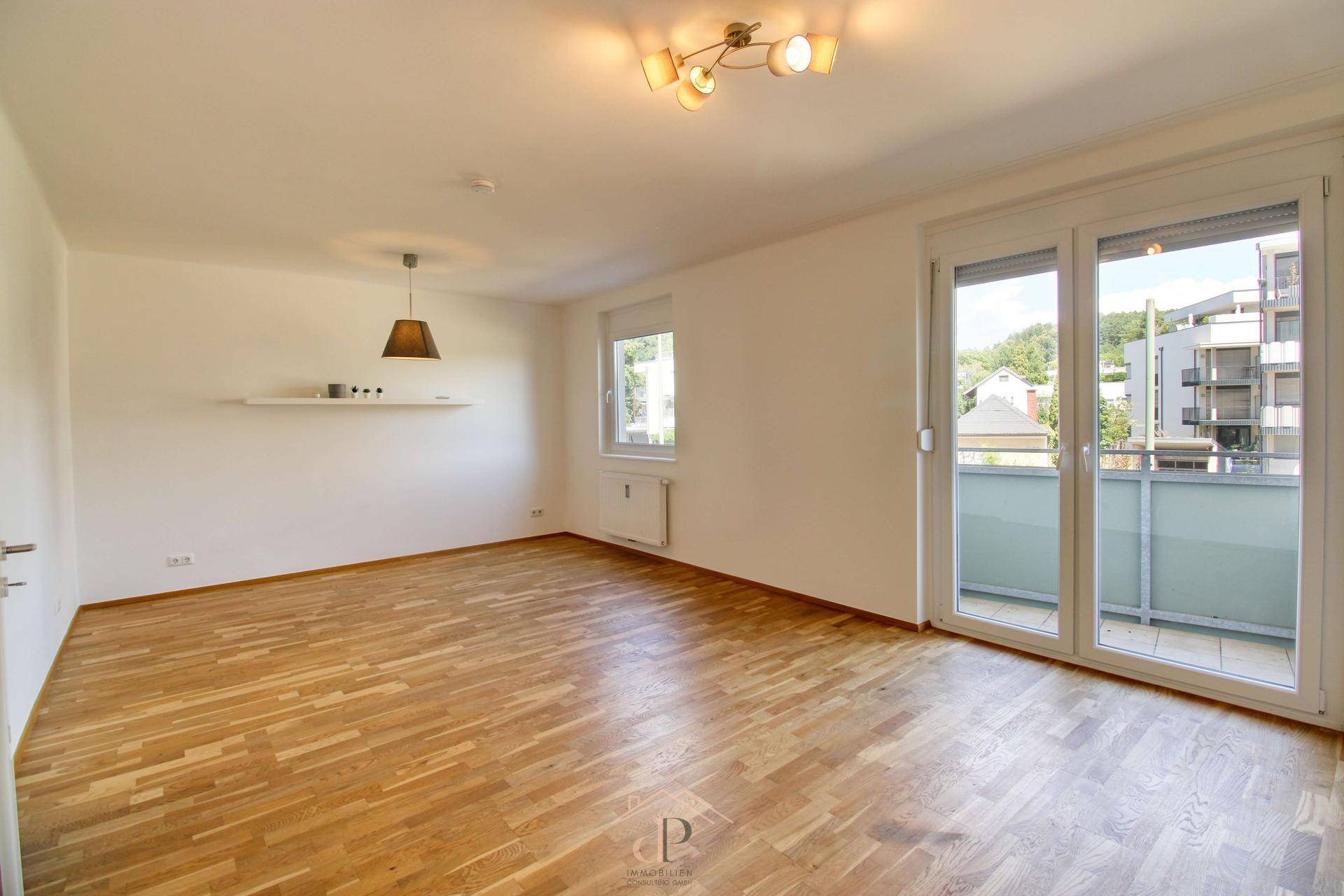 Geräumiges leeres Wohnzimmer mit hellem Parkettboden und Zugang zu einem Balkon.