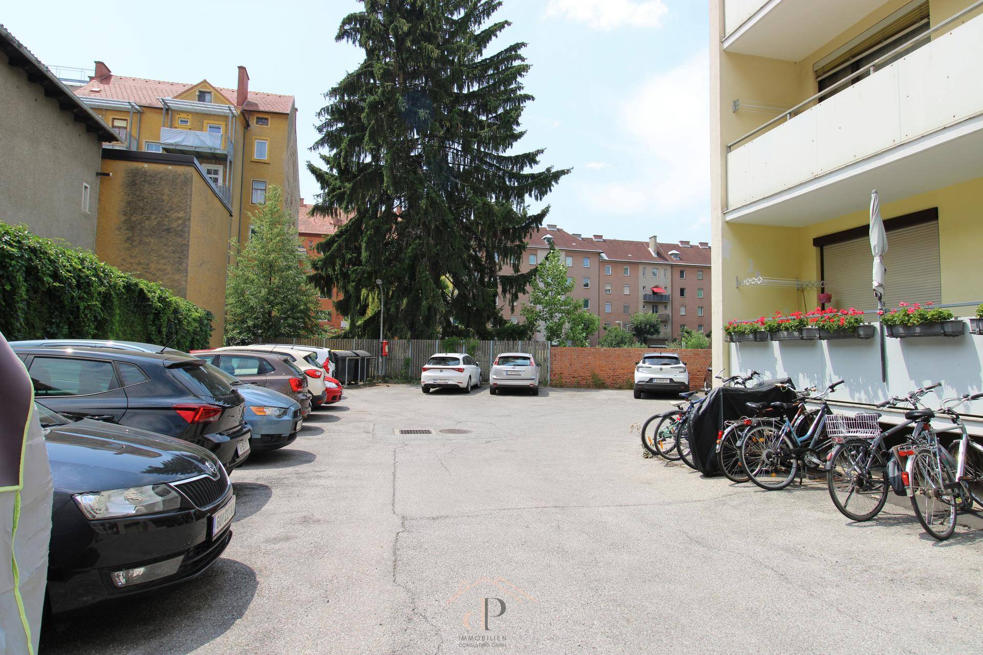 Außenparkplatz mit abgestellten Autos und Fahrrädern, umgeben von Wohngebäuden und Bäumen.