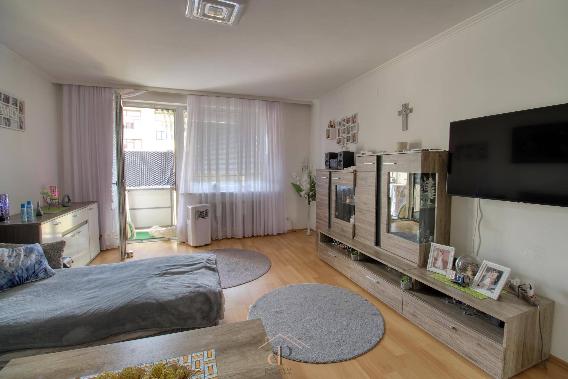 Gemütliches Wohnzimmer mit Sofa, Fernseher und Zugang zu einem Balkon, ausgestattet mit Holzmöbeln.