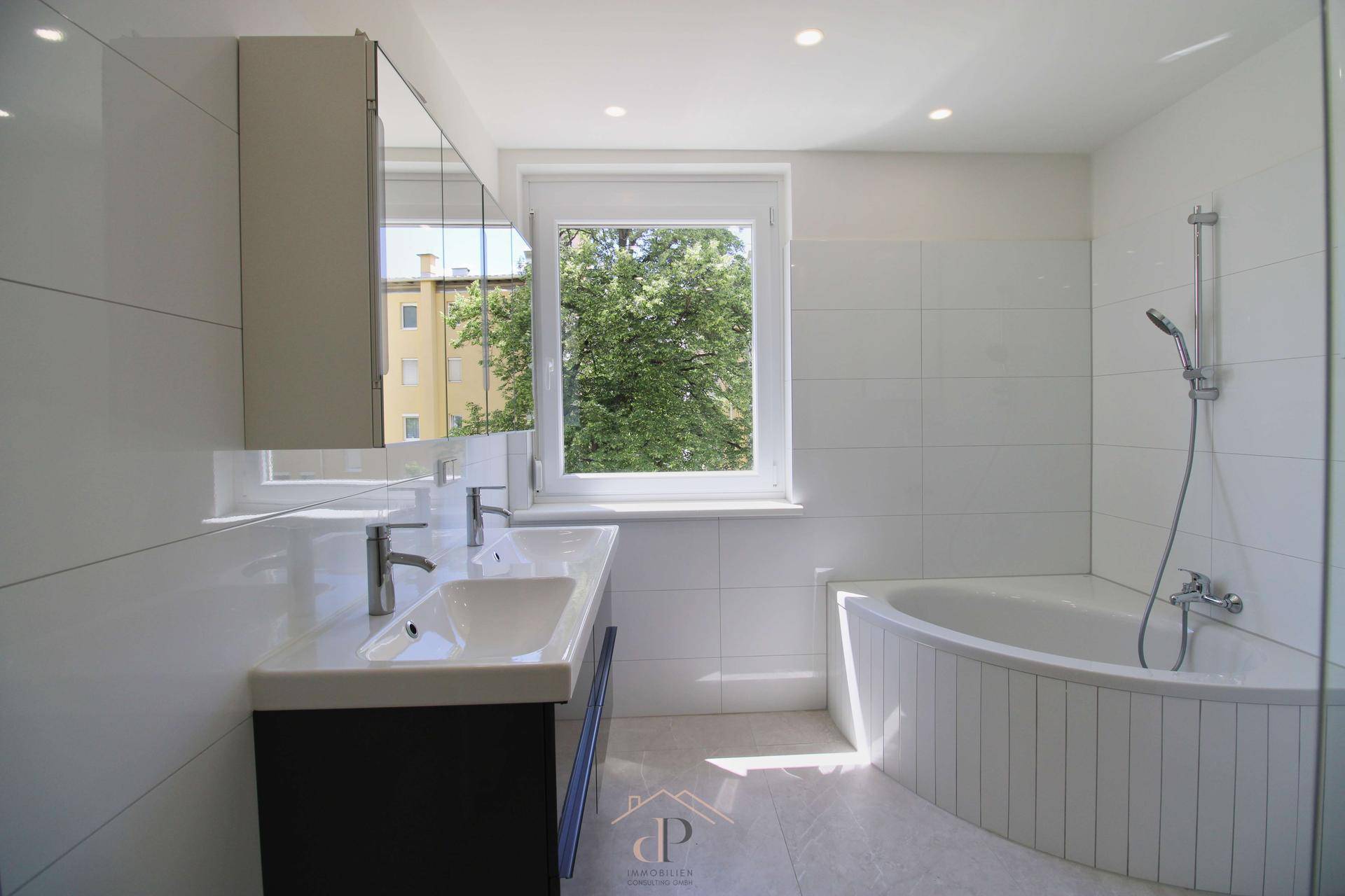 Helles Badezimmer mit Doppelwaschbecken, Badewanne und Fenster mit Blick ins Grüne.