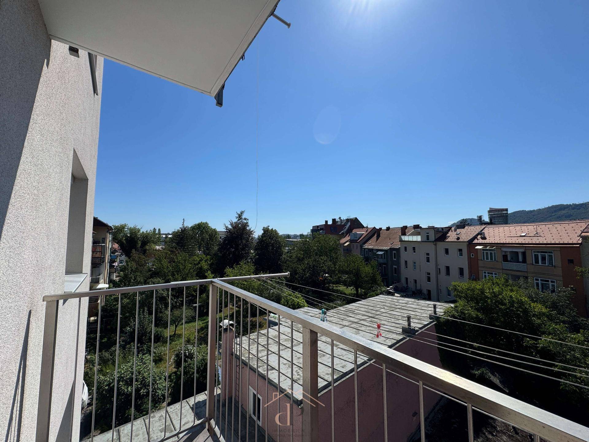 Sonniger Balkon mit Metallgeländer und Blick auf die umliegenden Gebäude und grüne Bäume.