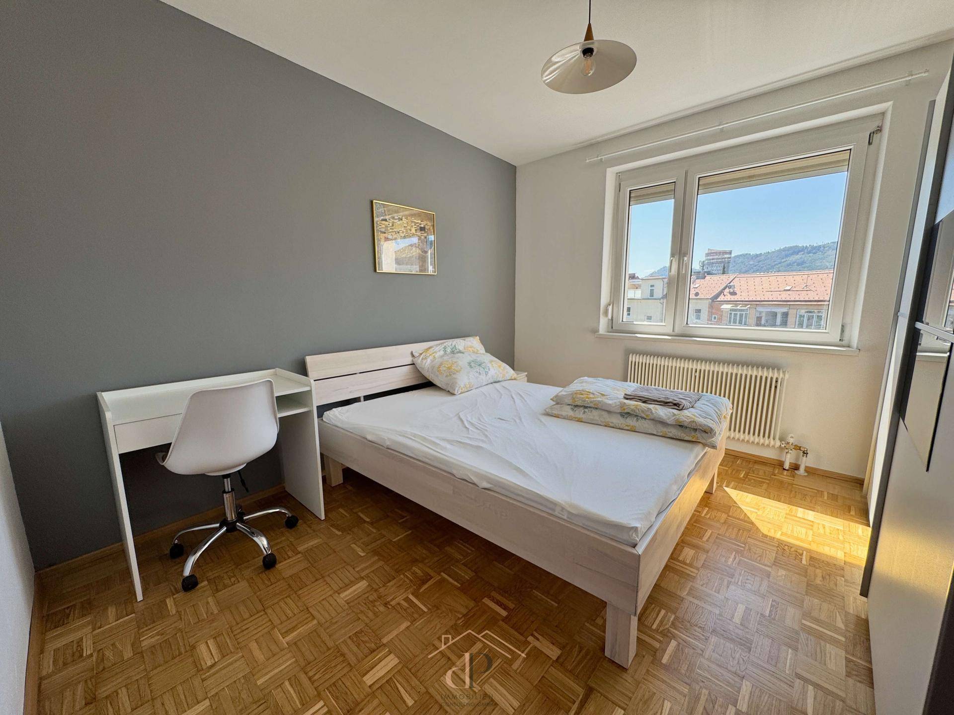 Helles Schlafzimmer mit Parkettboden, einem Bett und einem Schreibtisch vor einer grauen Wand.