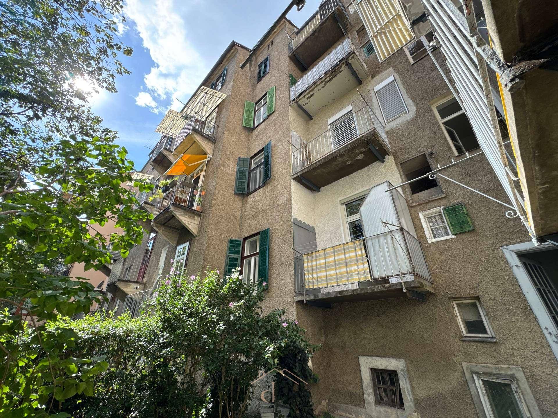 Historisches Gebäude mit mehreren Balkonen und grünen Fensterläden, umgeben von Bäumen.