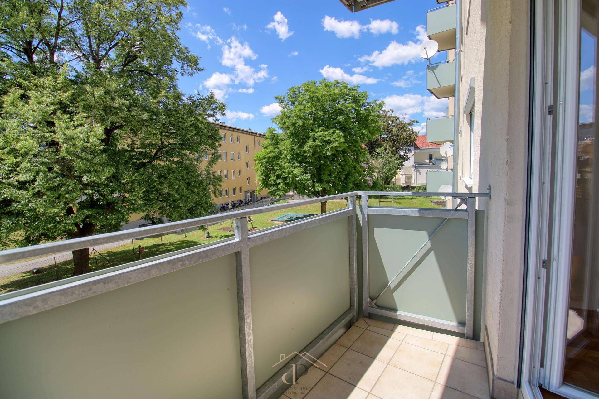Heller Balkon mit Metallgeländer und Blick auf grüne Bäume und umliegende Gebäude.