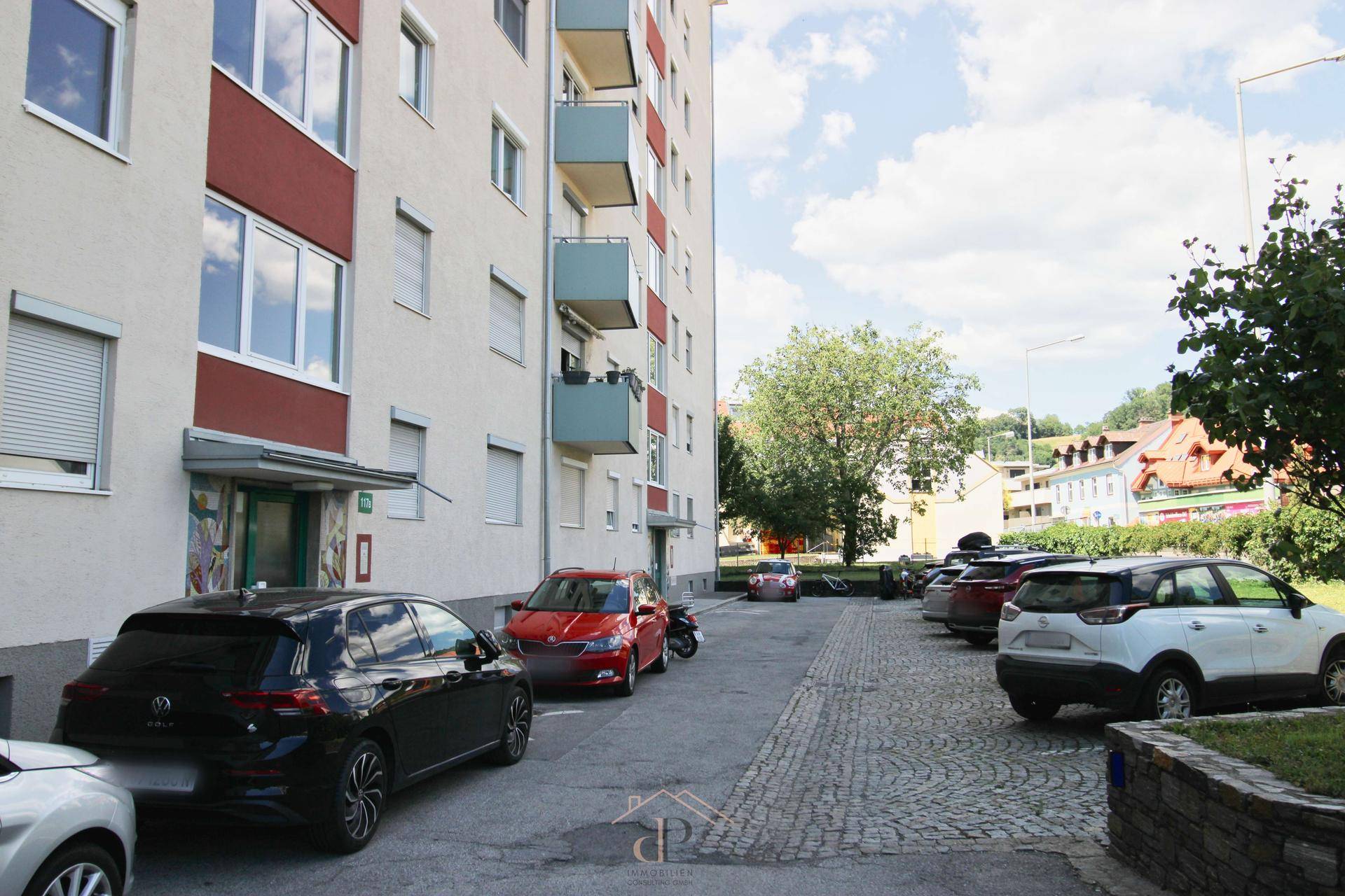 Außenansicht eines Wohngebäudes mit Parkplätzen und Bäumen an einer Straße.