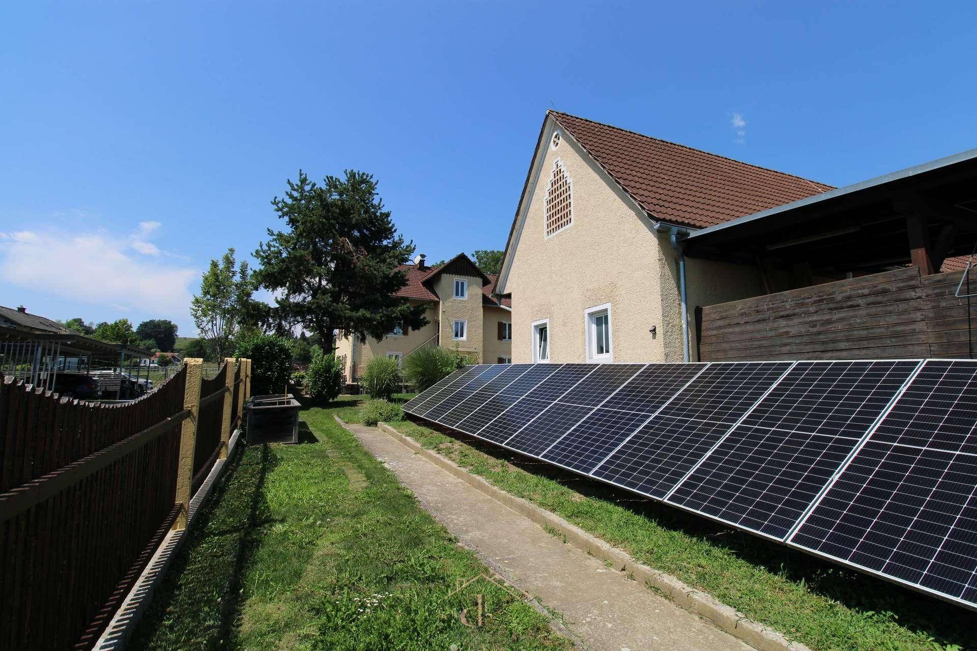 Gebäudeansicht mit Photovoltaikanlage im Vordergrund, umgeben von ländlicher Idylle und viel Grün.