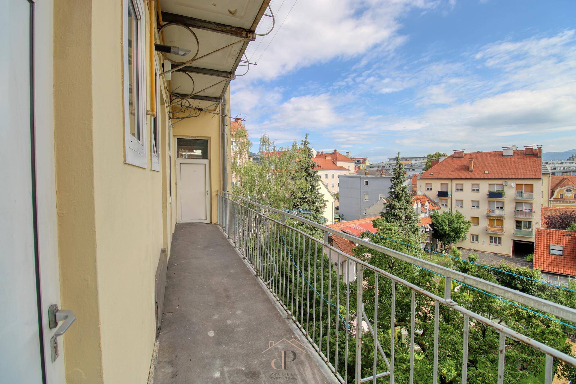 Langer Balkon mit Metallgeländer und Ausblick auf die umliegenden Stadtgebäude und Bäume.