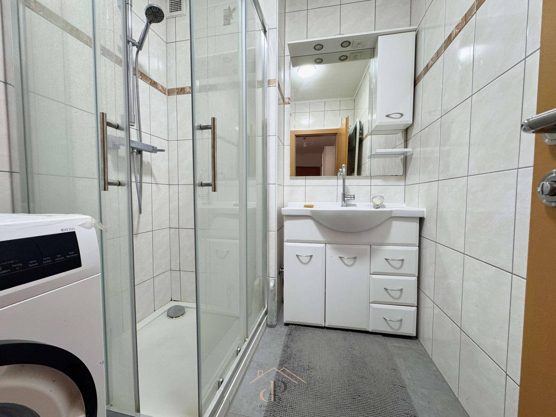 Modernes Badezimmer mit Duschkabine, Waschbecken und Waschmaschine in hellen Fliesen.