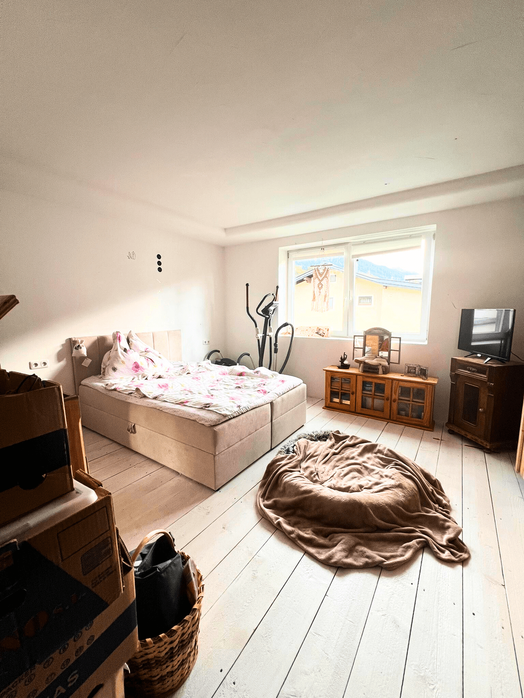 Schlafzimmer mit Bett, weißem Holzboden und einem Fenster, das viel Licht hereinlässt.
