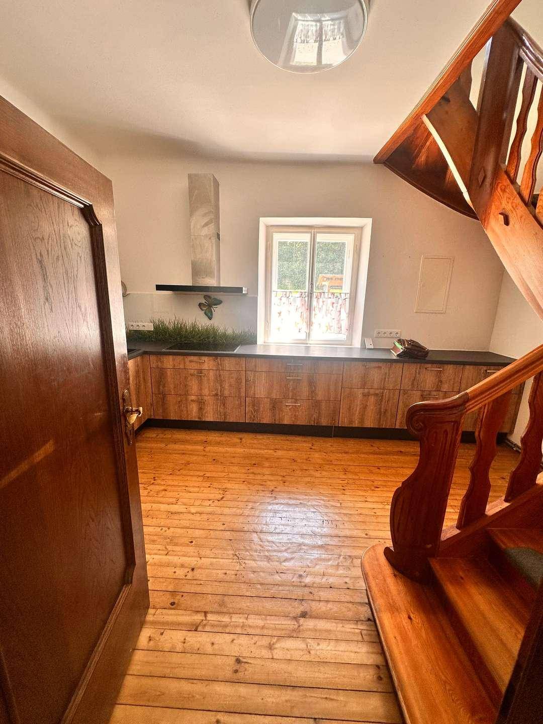 Küche mit Holzschränken, dunkler Arbeitsplatte und Dunstabzugshaube, daneben eine Holztreppe.