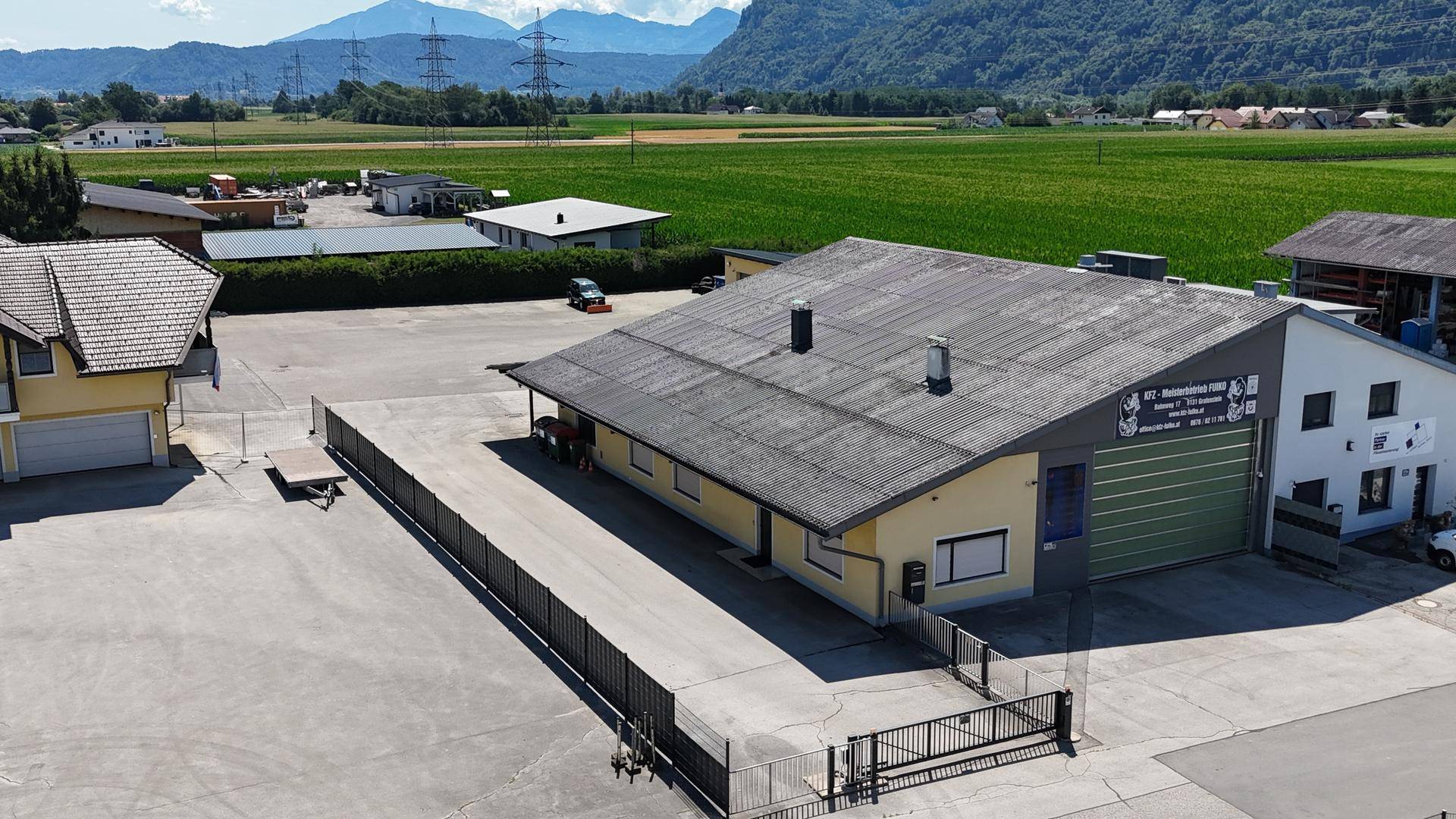 Gewerbegebäude mit großem Parkplatz und Blick auf die umliegende ländliche Landschaft und die Berge im Hintergrund.