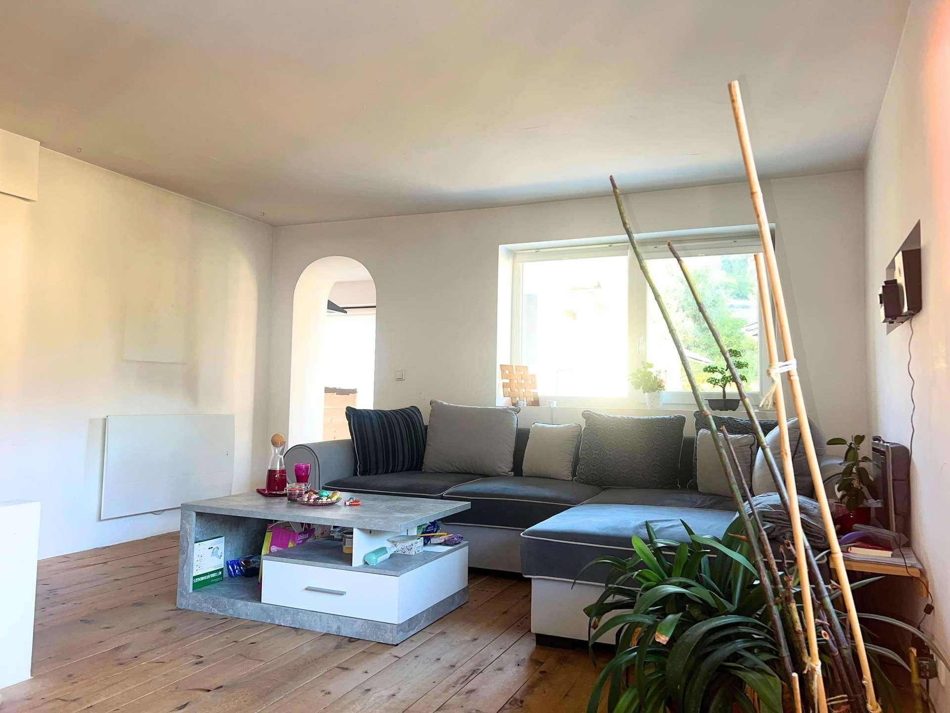 Wohnzimmer mit grauem Ecksofa, Couchtisch und Holzboden, mit einem Durchgang zu einem weiteren Raum.