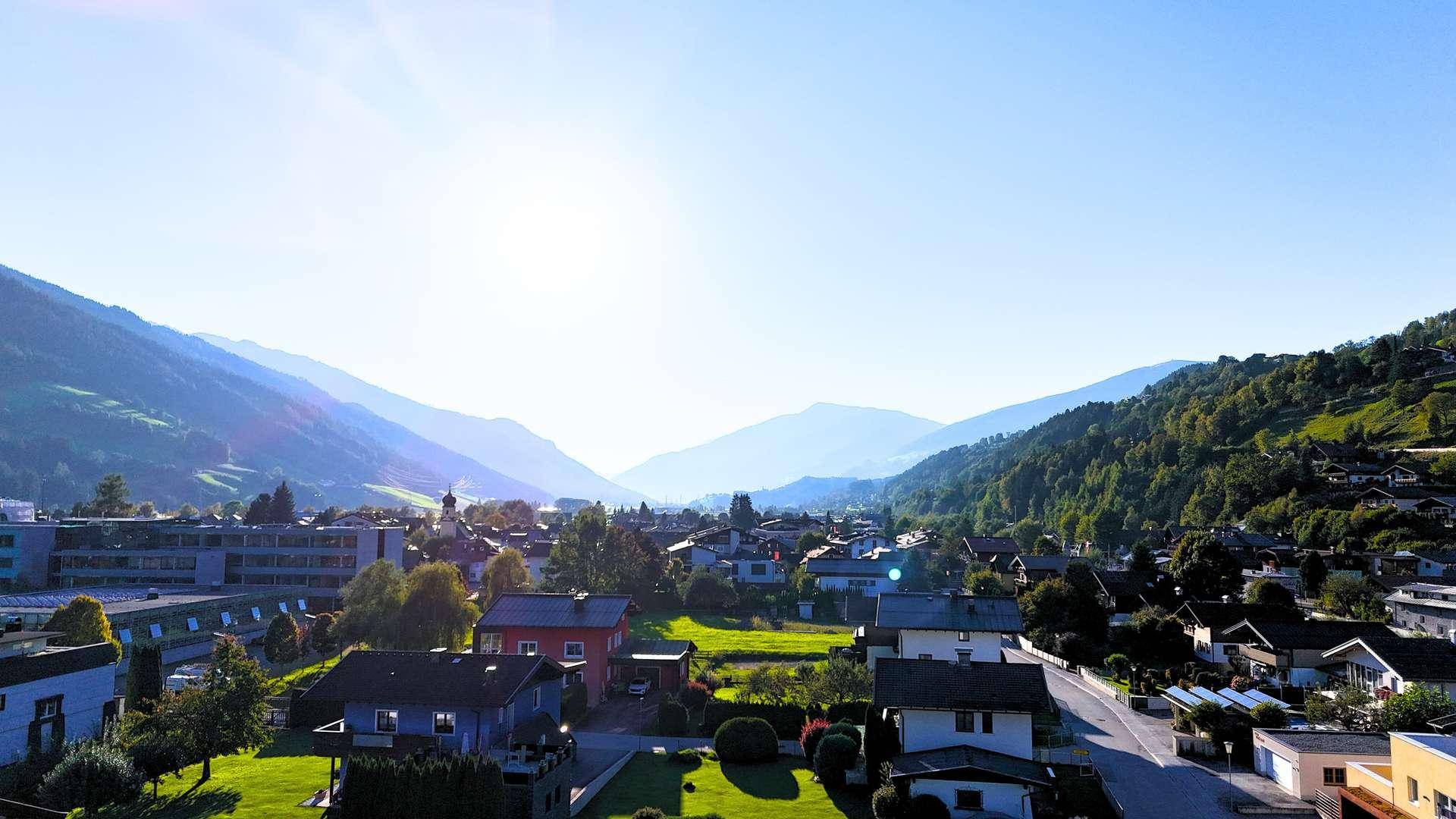 Sonniger Ausblick auf eine Siedlung im Tal, umgeben von grünen Bergen.