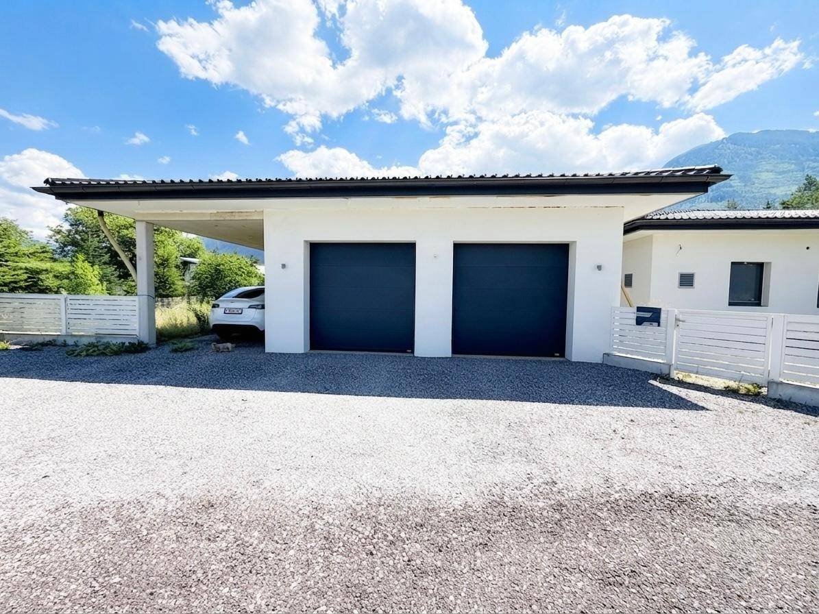 Modernes Gebäude mit Doppelgarage und Carport, umgeben von einer Schotterauffahrt und Bergen.